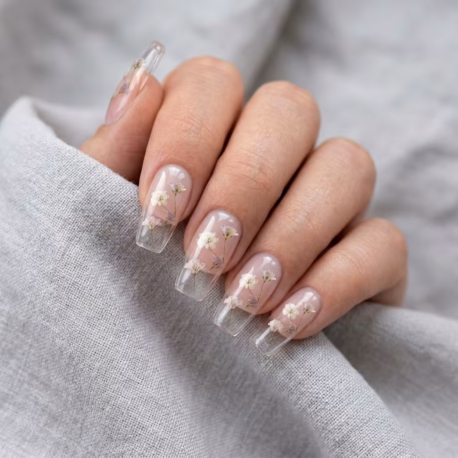 Mão com cinco unhas quadradas no Gel Construtor Cristal Clear, transparente, com flores secas encapsuladas, sobre linho cinza