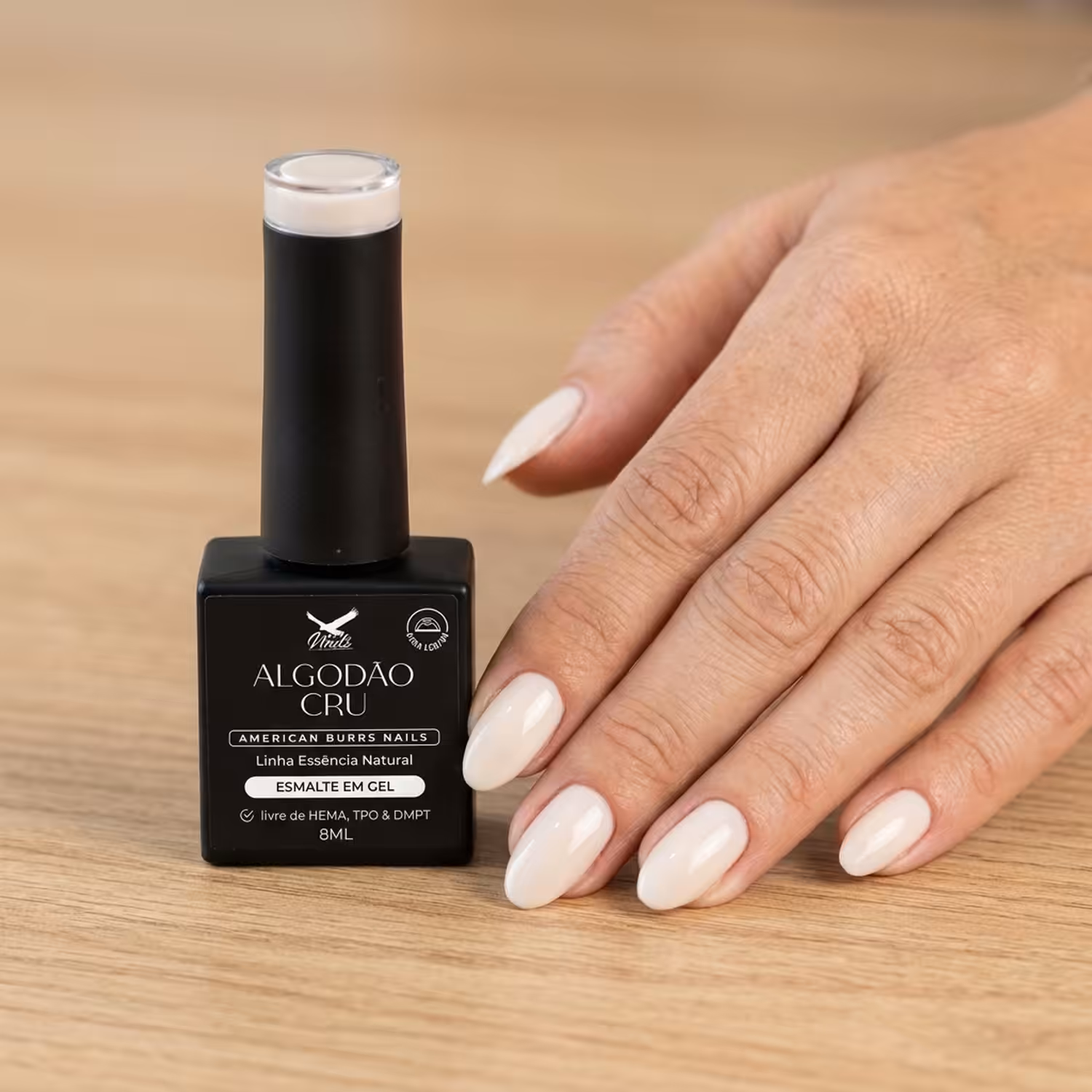 Frasco do Esmalte em Gel Algodão Cru ao lado de uma mão apoiada na mesa, com as unhas pintadas na cor
