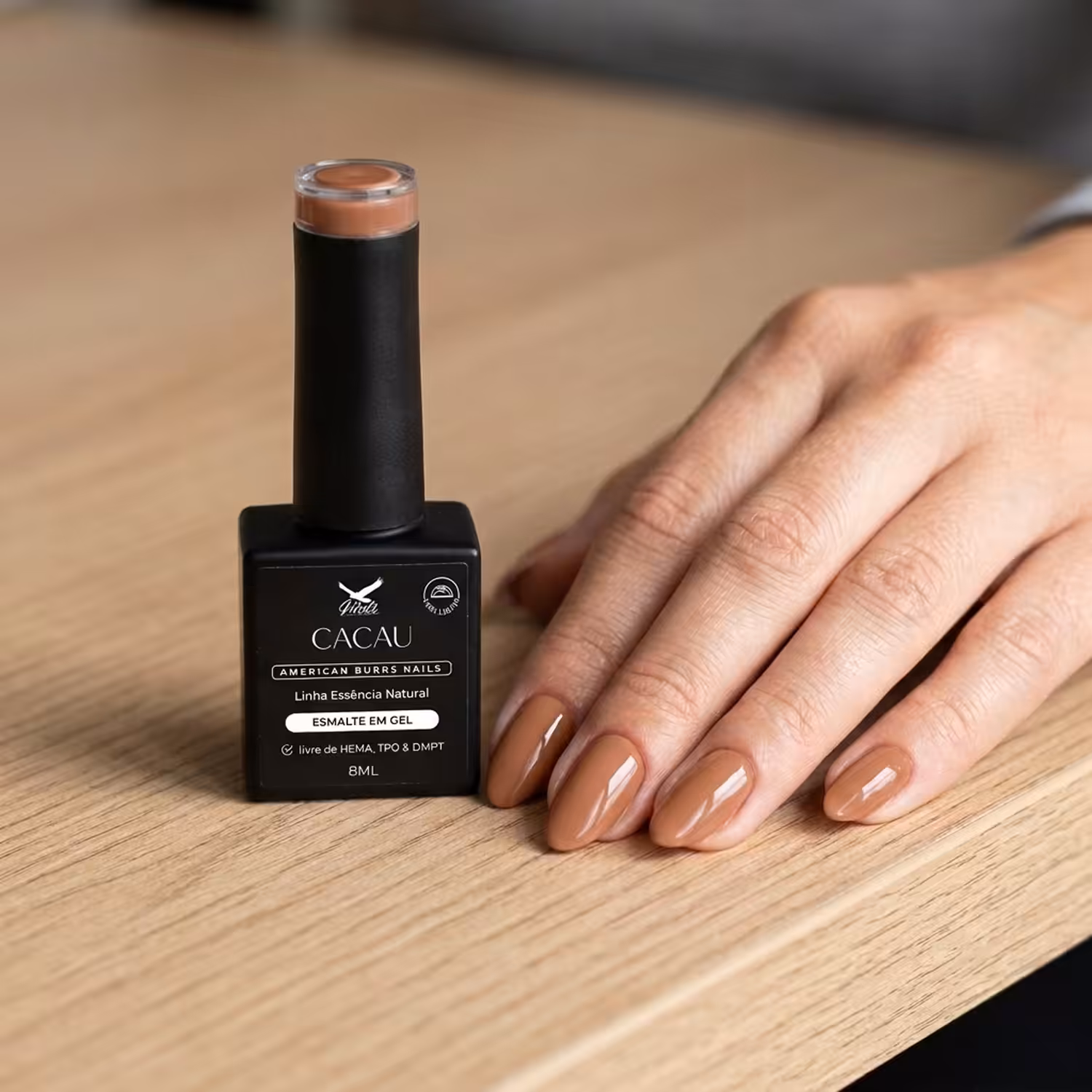 Frasco do Esmalte em Gel Cacau ao lado de uma mão apoiada na mesa, com as unhas pintadas na cor