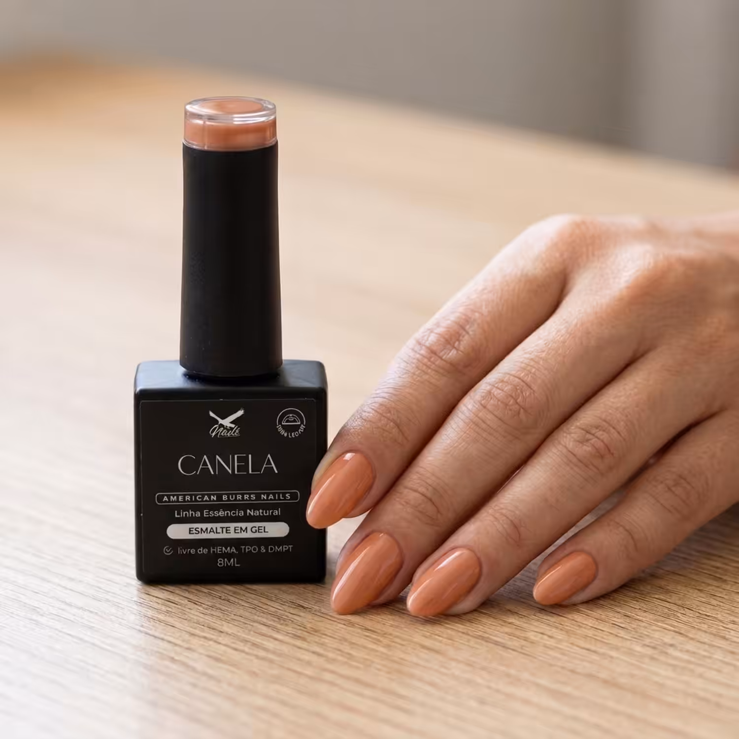 Frasco do Esmalte em Gel Canela ao lado de uma mão apoiada na mesa, com as unhas pintadas na cor