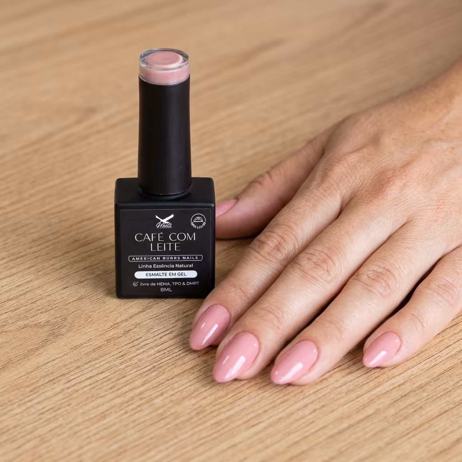 Frasco do Esmalte em Gel Café com Leite ao lado de uma mão apoiada na mesa, com as unhas pintadas na cor
