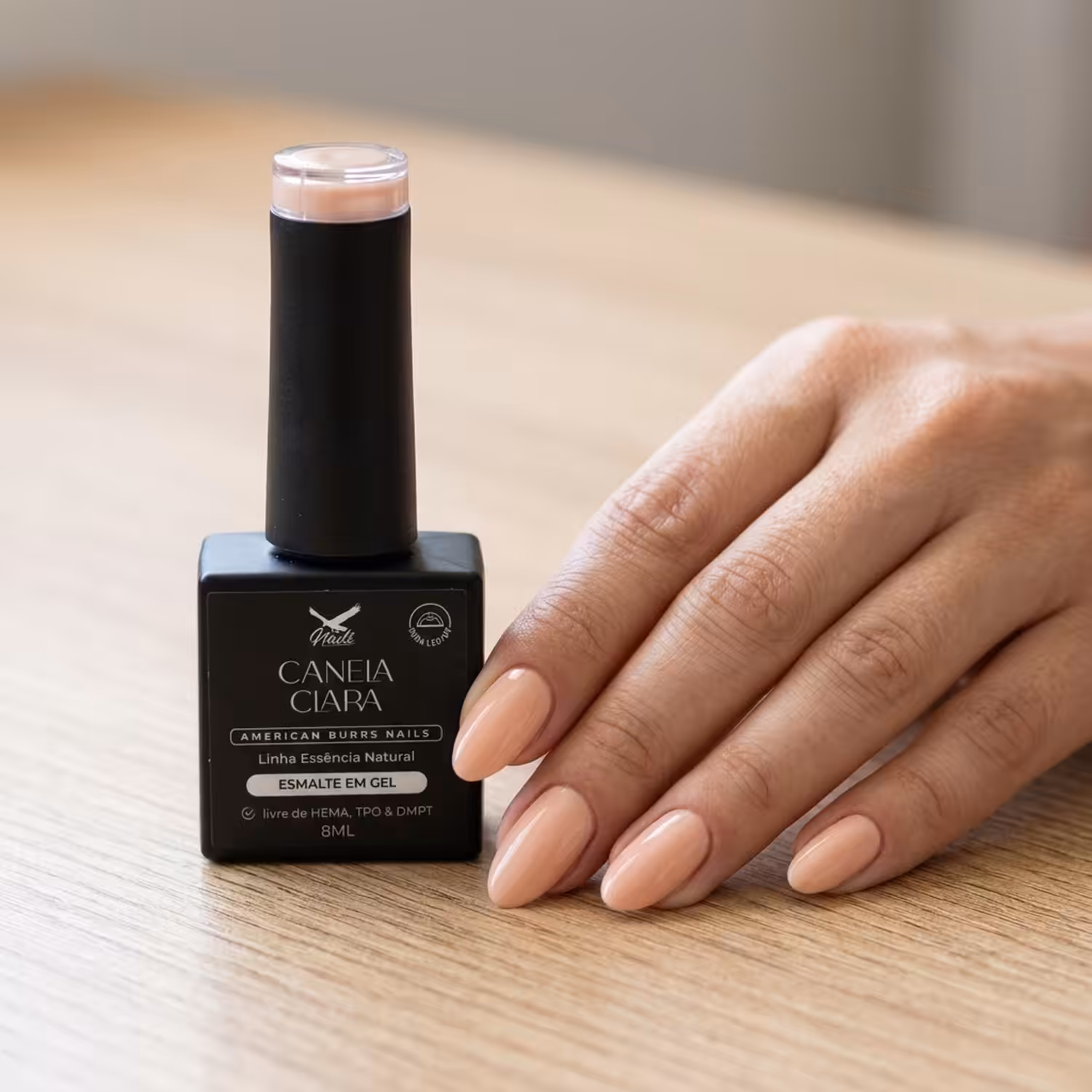 Frasco do Esmalte em Gel Canela Clara ao lado de uma mão apoiada na mesa, com as unhas pintadas na cor