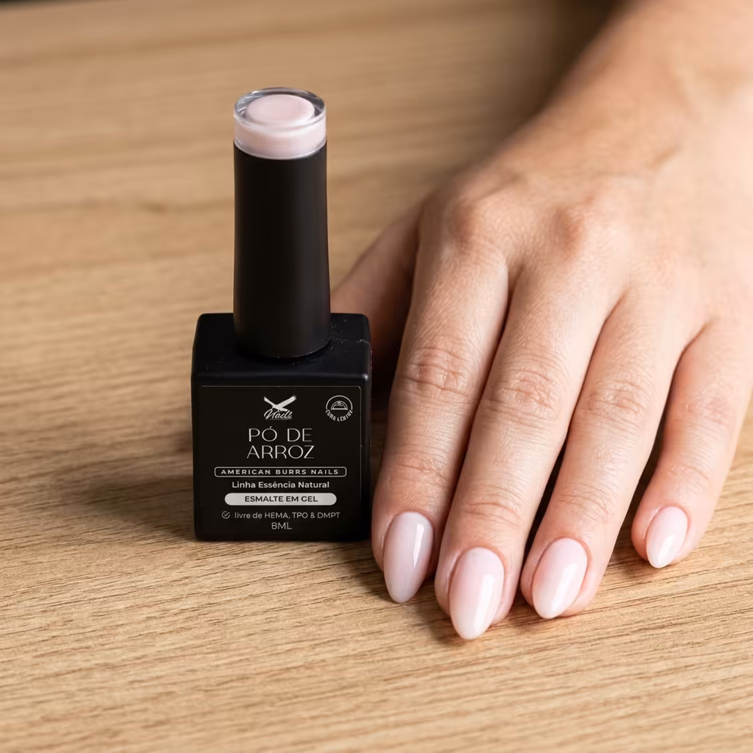 Frasco do Esmalte em Gel Pó de Arroz ao lado de uma mão apoiada na mesa, com as unhas pintadas na cor