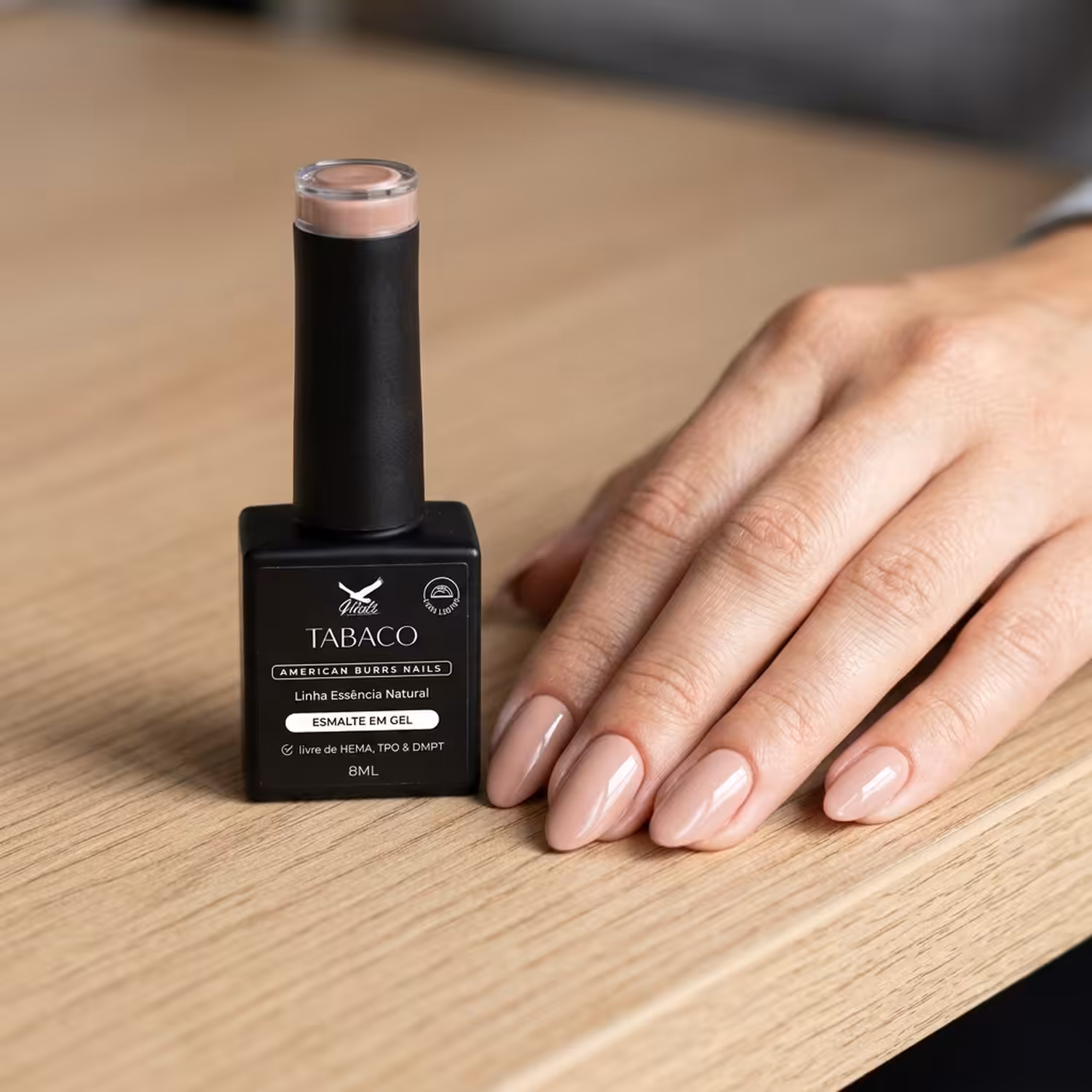 Frasco do Esmalte em Gel Tabaco ao lado de uma mão apoiada na mesa, com as unhas pintadas na cor